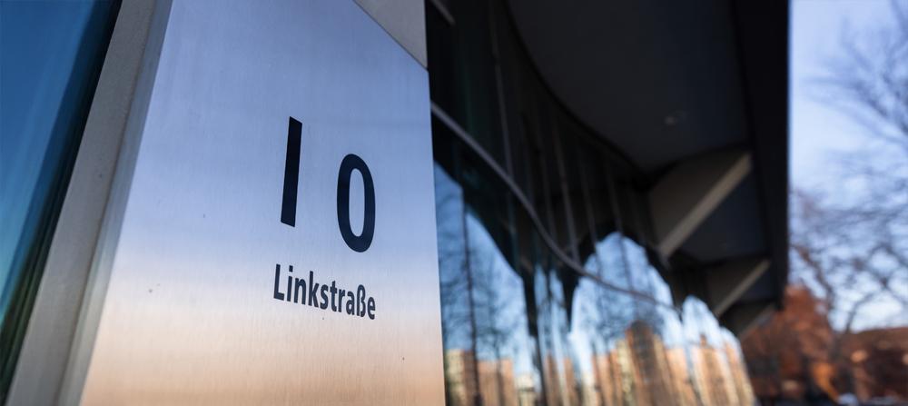  Nahaufnahme eines glänzenden Metallschilds an einem Gebäude, auf dem in schwarzer Schrift die Hausnummer "10" und darunter "Linkstraße" steht. Rechts spiegelt sich die Umgebung in der gebogenen Glasfassade des Gebäudes, im Hintergrund sind Bäume und ein blauer Himmel sichtbar.